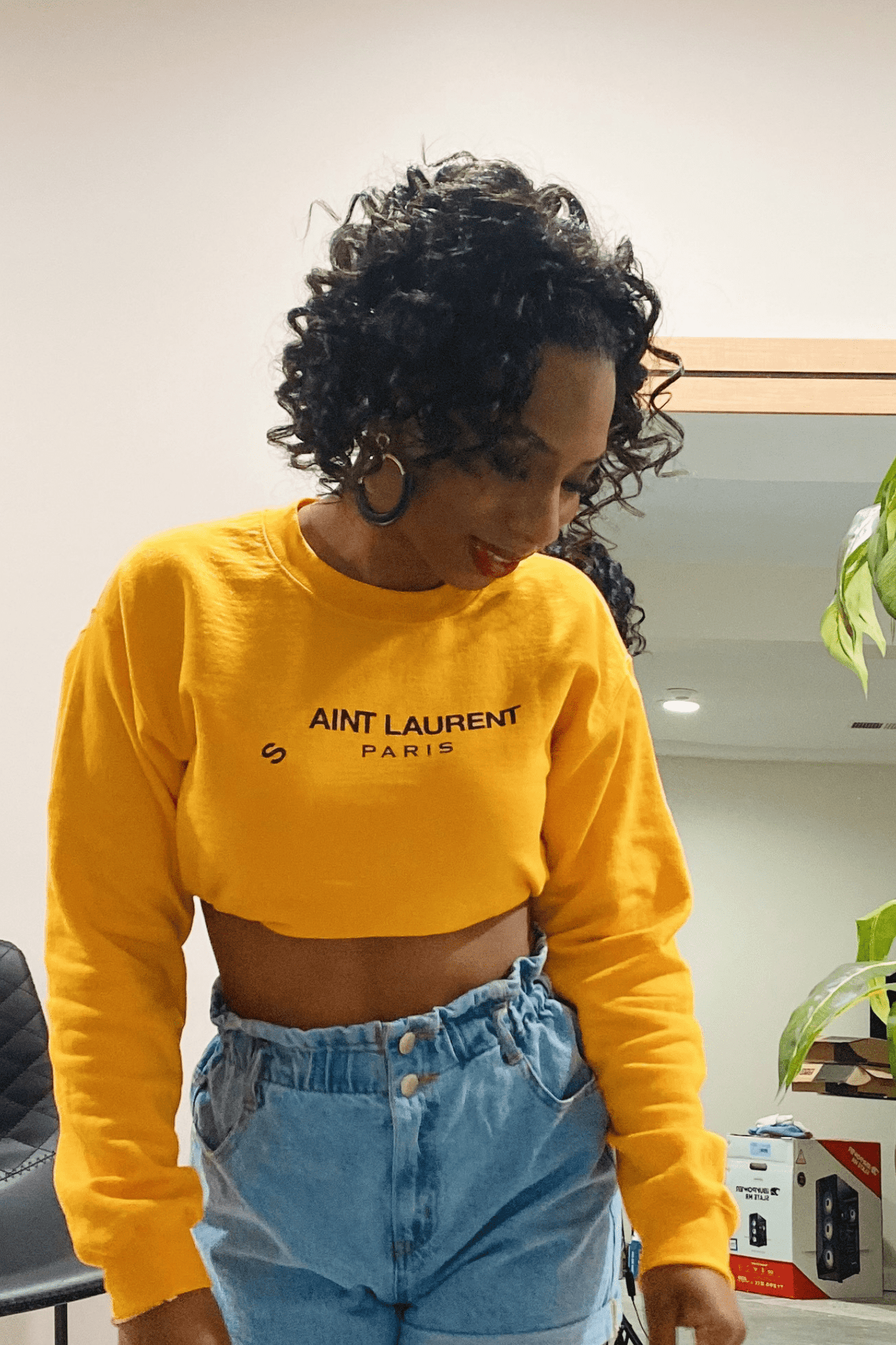 Women's yellow long sleeve crop sweatshirt that has "Saint Laurent Paris" written in bold black print on the front pained with denim shorts.