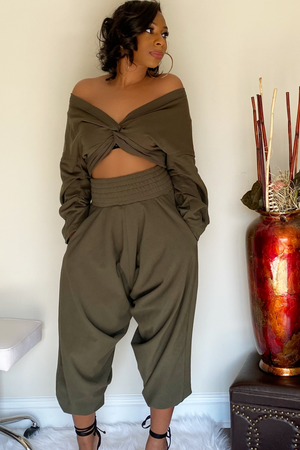 Front view of the olive-green harem pants with the long sleeve crop top. There is a knot design in the middle of the shirt. The model has both sleeves pulled down exposing the shoulders. The harem bottoms have a wide slightly ruffled waist band. The bottoms are wigged and stop around the ankle.