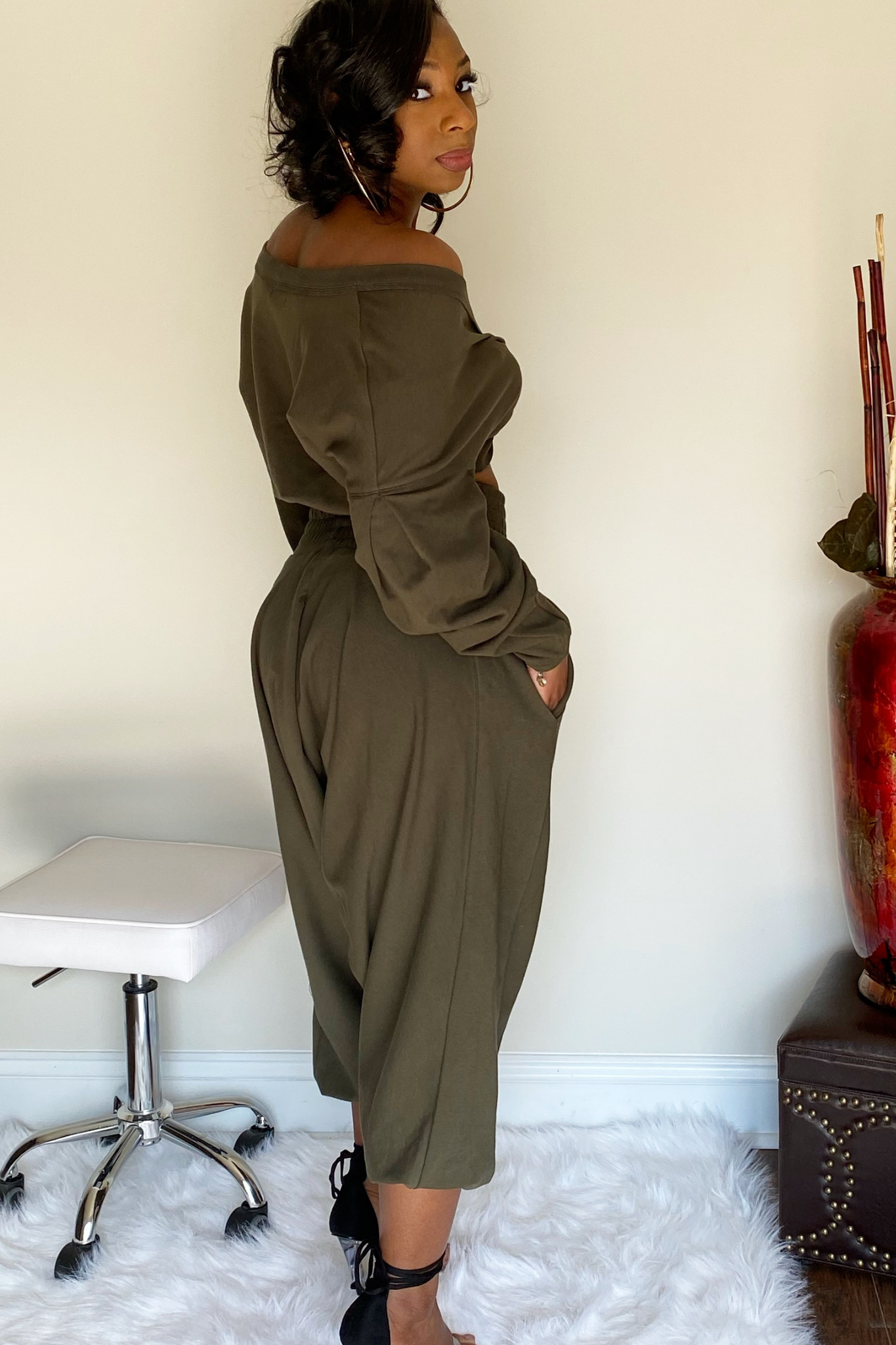 Back view of the olive-green harem pants with the long sleeve crop top. There is a knot design in the middle of the crop shirt. The shirt is slightly pulled up to expose the model's midsection. The model has the sleeve down exposing the right shoulder. The harem bottoms have a wide slightly ruffled waist band. The bottoms are wide legged and stop around the ankle. Model has her hand in the right pocket.