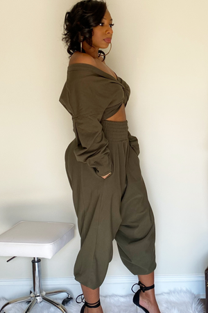 Side view of the olive-green harem pants with the long sleeve crop top. There is a knot design in the middle of the crop shirt. The shirt is slightly pulled up to expose the model's midsection. The model has the sleeve down exposing the right shoulder. The harem bottoms have a wide slightly ruffled waist band. The bottoms are wide legged and stop around the ankle. Model has her hand in the right pocket.