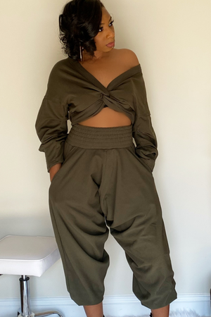 Front view of the olive-green harem pants with the long sleeve crop top. There is a knot design in the middle of the shirt. The model has the sleeve pulled down exposing the left shoulder. The harem bottoms have a wide slightly ruffled waist band. The bottoms are wide legged and stop around the ankle.