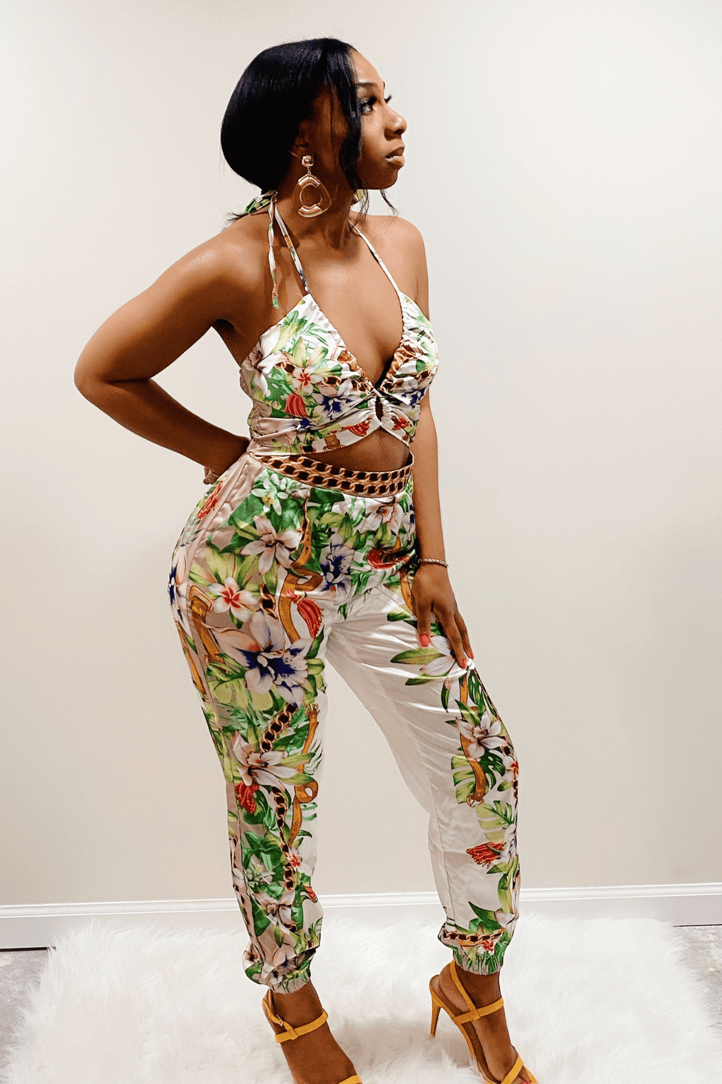 Front view of the white jumpsuit with flower pattern along the legs and top. The flower pattern is yellow, green, with areas of purple, pink and red. The floral design goes along the outside of the legs and covers the top. The jumpsuit has a triangle shaped stomach cutout above the chain designed waist. The top of the jumpsuit has a halter design that ties around the neck.