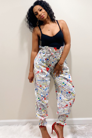 Front view of the "Read All About IT" Pants. Pants have a white background with an all over red, yellow, blue and black paint splatter design. The waist has a paperback design with a matching belt. At the bottom of the pants there are attached ankle straps to tie for a jogger pants style look. Model paired the pants with a solid black tank top.