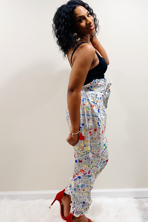 Side view of the "Read All About IT" Pants. Pants have a white background with an all over red, yellow, blue and black paint splatter design. The waist has a paperback design with a matching belt. At the bottom of the pants there are attached ankle straps to tie for a jogger pants style look. Model paired the pants with a solid black tank top.