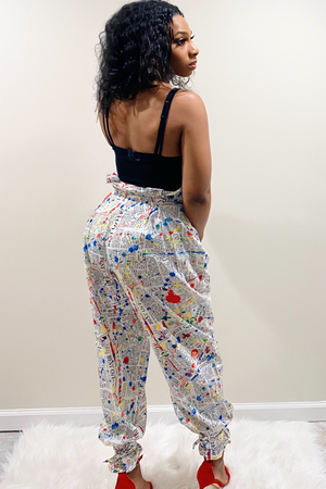 Back view of the "Read All About IT" Pants. Pants have a white background with an all over red, yellow, blue and black paint splatter design. The waist has a paperback design with a matching belt. At the bottom of the pants there are attached ankle straps to tie for a jogger pants style look. Model paired the pants with a solid black tank top.