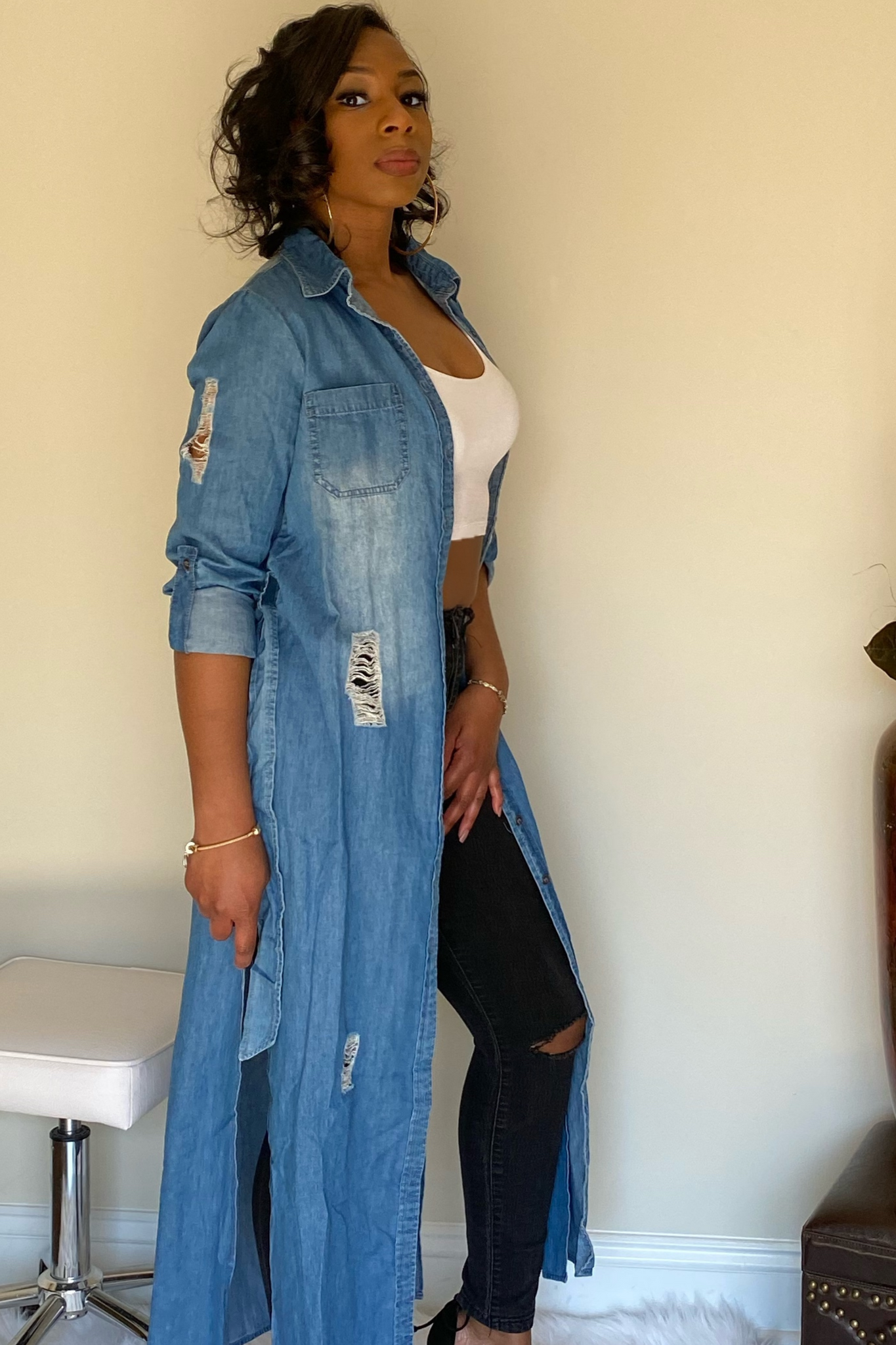 Side view of open denim shirt that displays a white crop t-shirt and black jeans underneath the shirt. The denim shirt has a collar along with buttons from the collar down to the calf area. The shirt has a slit along the bottom that starts toward the bottom of the thigh that goes all the way through to the bottom of the shirt on both sides.